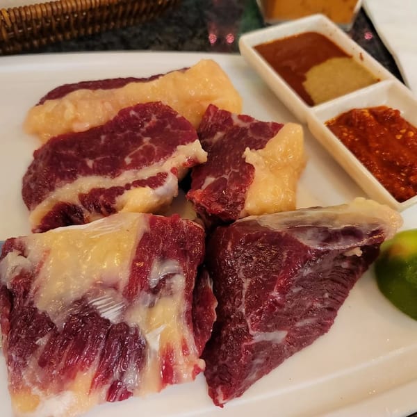 Raw beef cubes with fat, lime, and three dipping sauces on a white plate.
