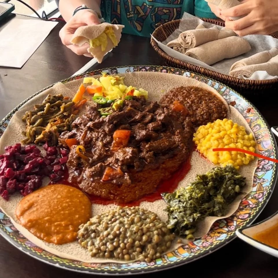 An Ethiopian injera platter with various stews, vegetables, and lentils, with a hand holding injera.